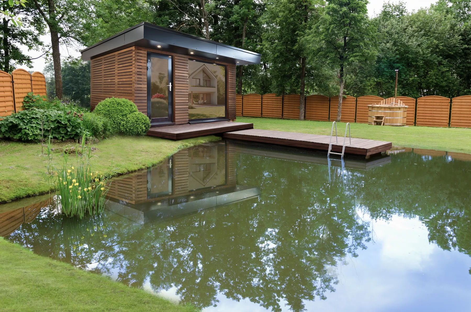Außensauna im Garten am Schwimmteich: ein Kubus mit Flachdach und großen Glasflächen, davor ein Holzdeck mit Badeleiter, im Wasser spiegelt sich die Kabine.